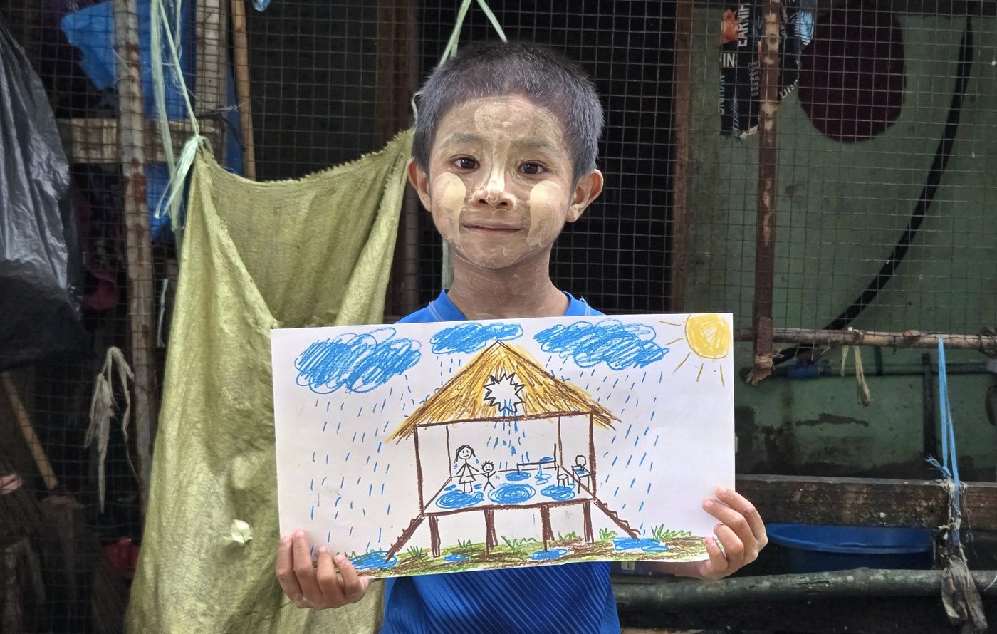A boy in rural Myanmar with his drawing of his family's home, showing rain leaking through the roof